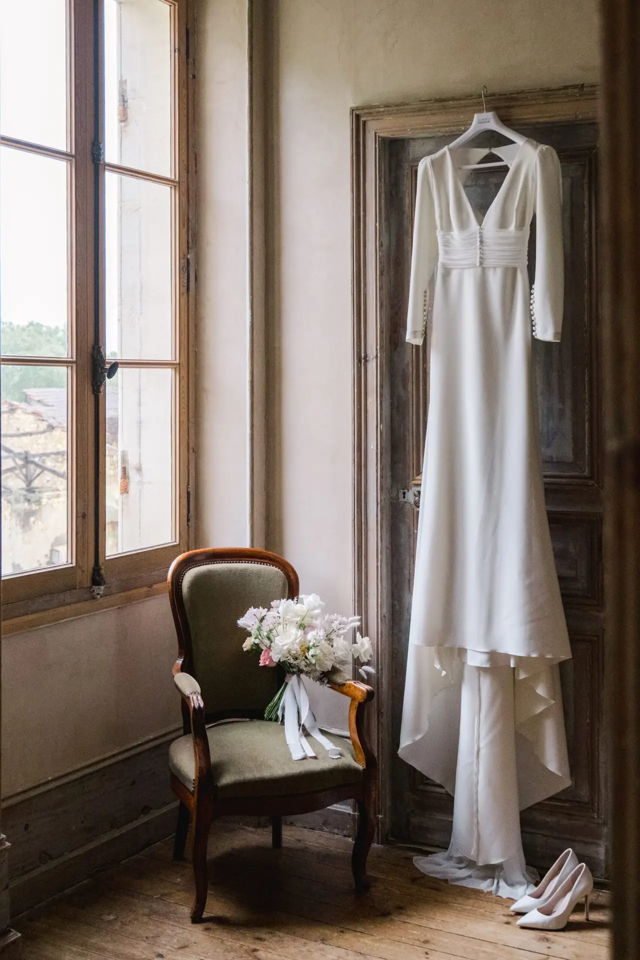 Minimalist wedding dress hanging in a rustic bridal suite at Château Fengari, alongside the bouquet and heels.