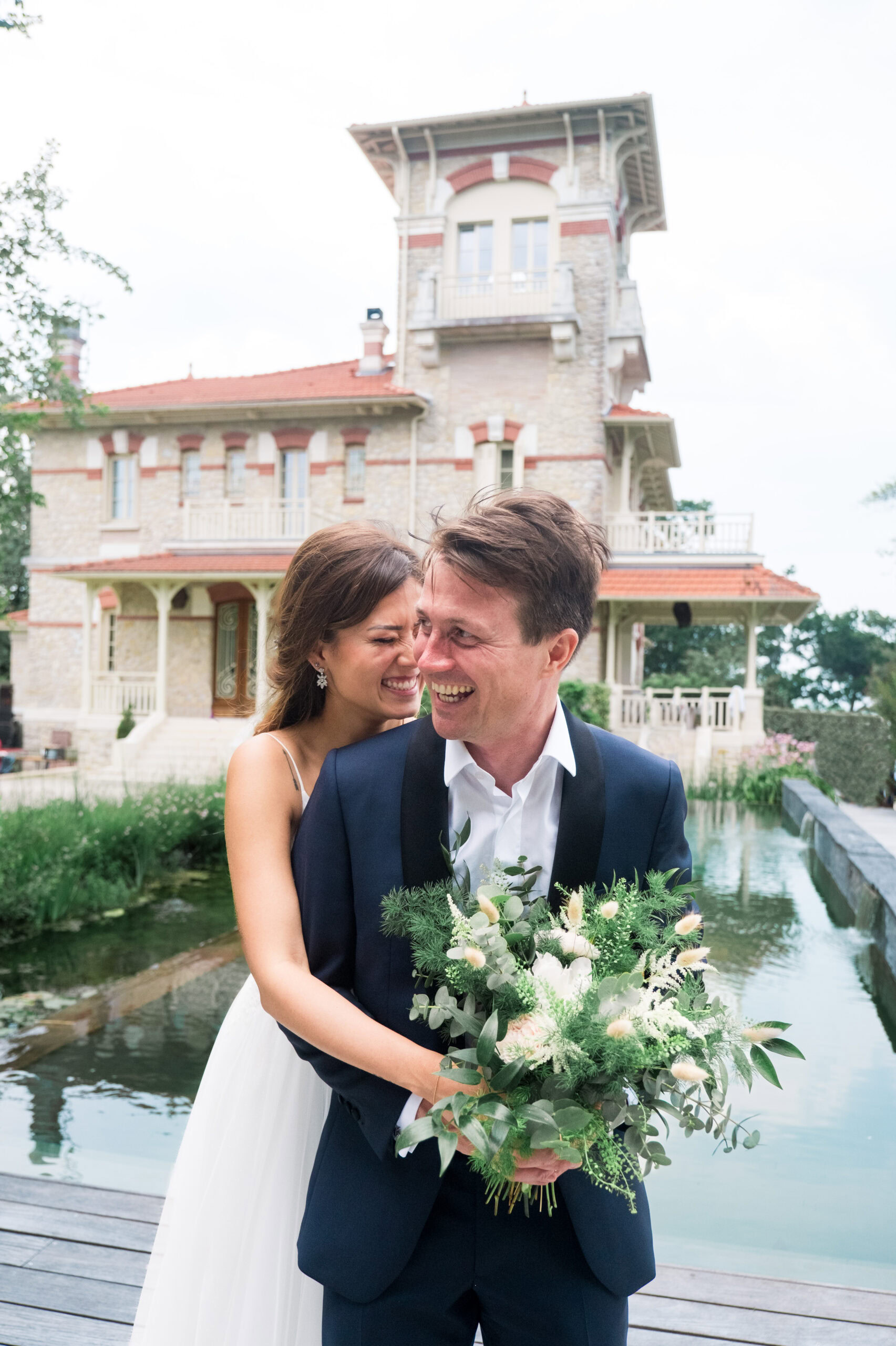 Fous rires des mariés devant une villa en pierres au bord de l’eau, scène pleine de spontanéité et d’amour.