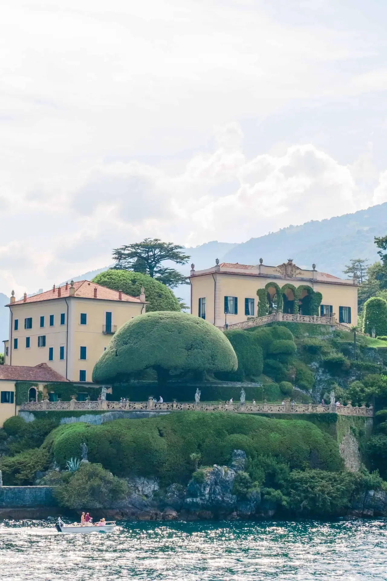 Scenic view of Villa Balbianello perched on a cliff above Lake Como, with manicured gardens, historic yellow buildings, and dramatic trees under a cloudy sky, captured from a boat on the water