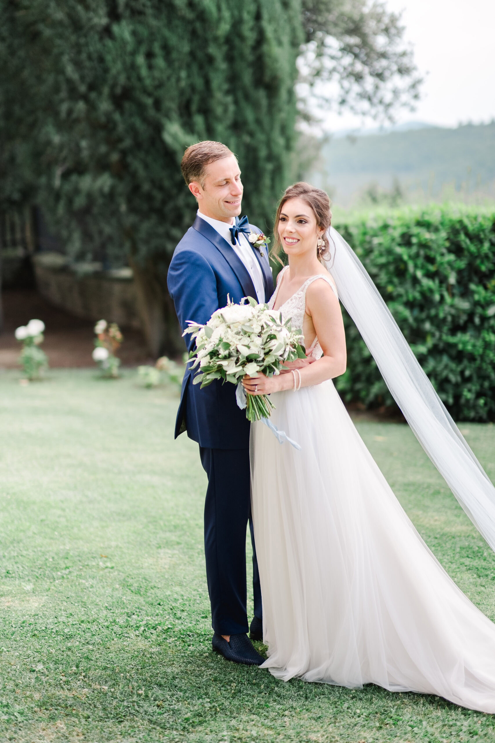 Jeunes mariés dans un jardin toscan, le voile de la mariée flottant dans la lumière douce d’un après-midi en Italie.