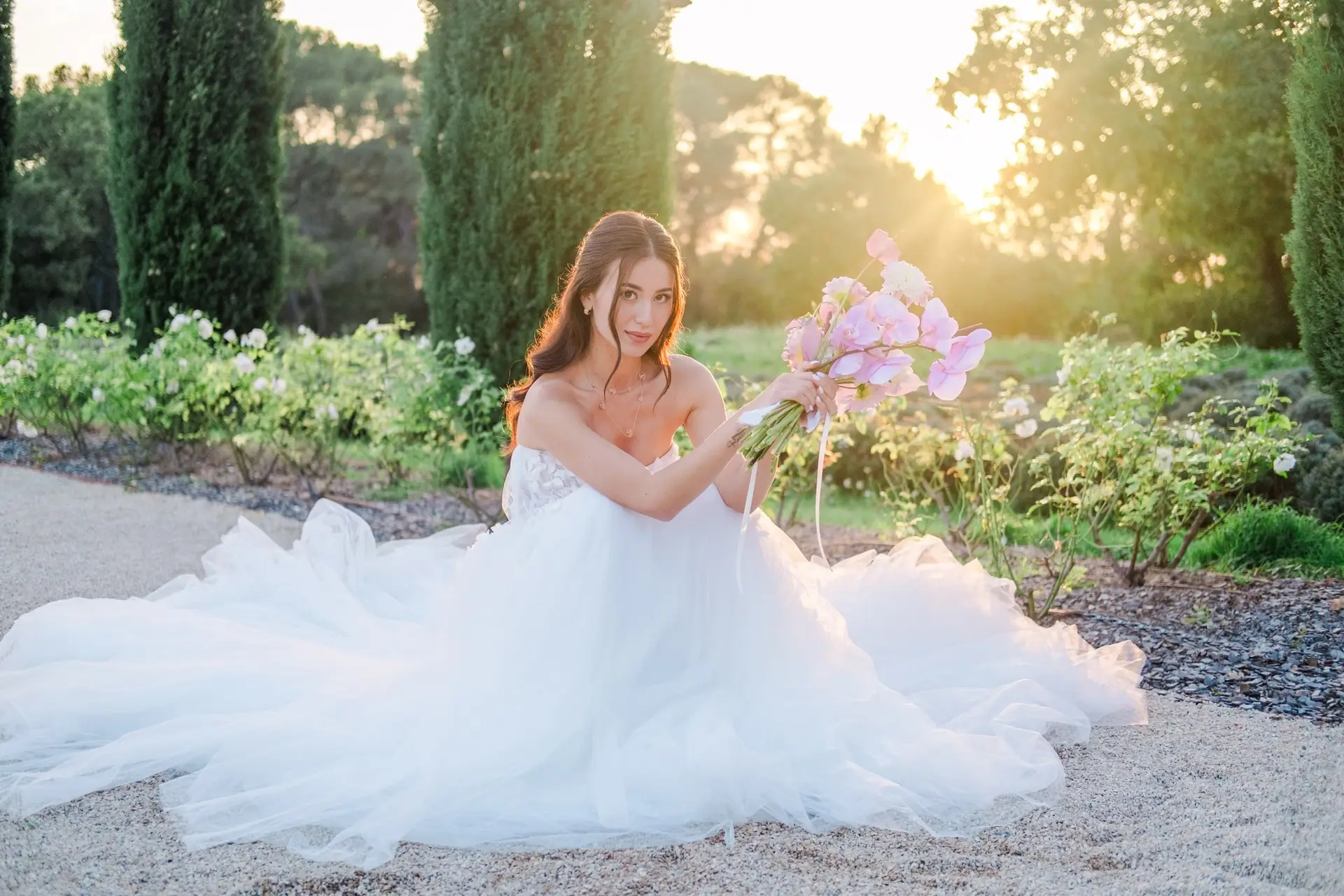 Bride sitting on the garden path with her gown spread around her, holding a bouquet at sunset