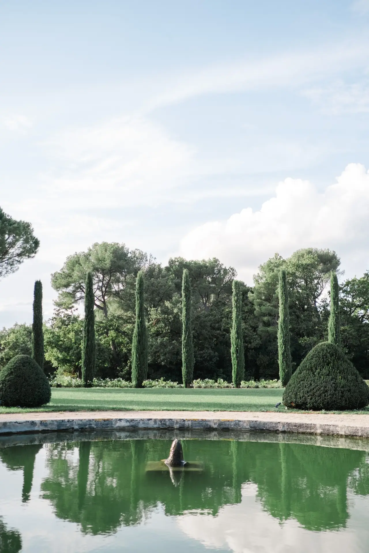 Pond with a fountain surrounded by manicured hedges and tall cypress trees at Château de la Gaude.