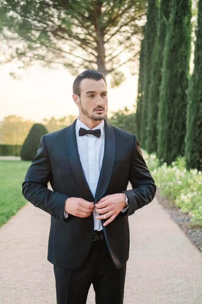 Groom adjusting his tuxedo jacket while standing on a garden path at Château de la Gaude
