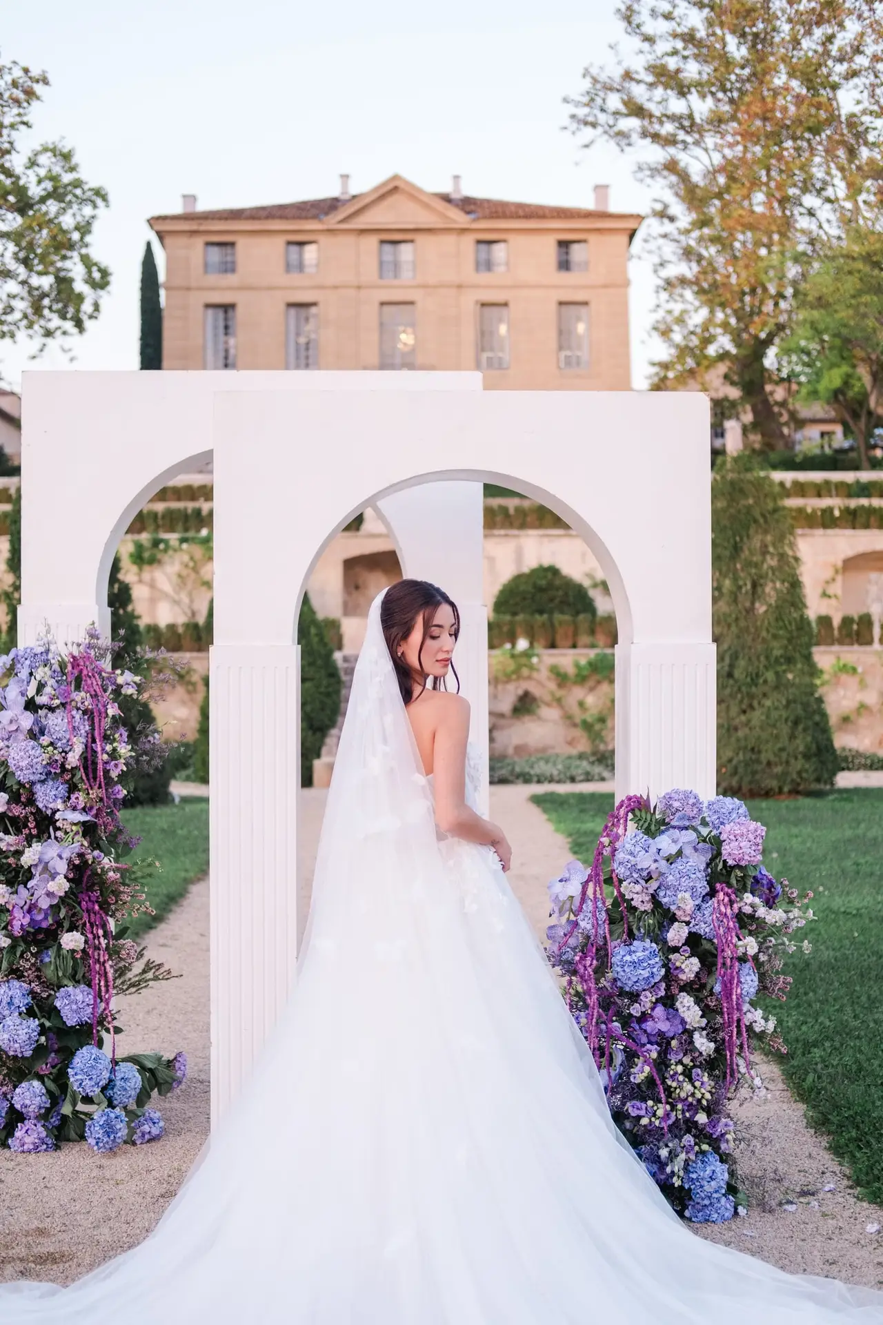 Bride standing under a white ceremony arch with purple floral arrangements at Château de la Gaude