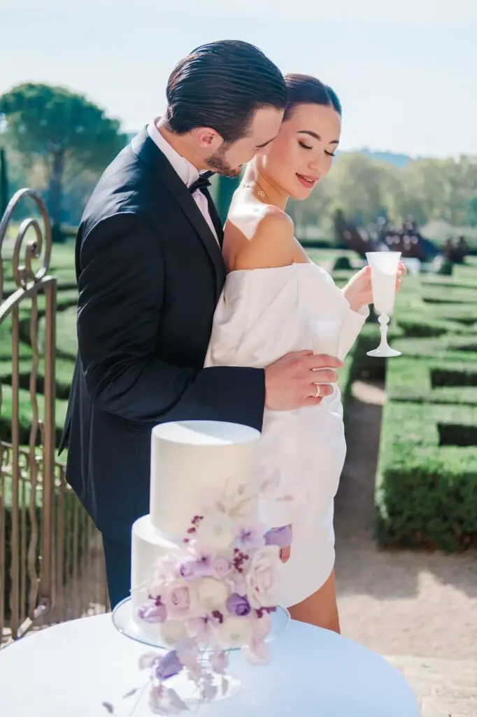 Elegant bride and groom enjoying champagne by a white wedding cake adorned with pastel flowers in the gardens of Château de la Gaude