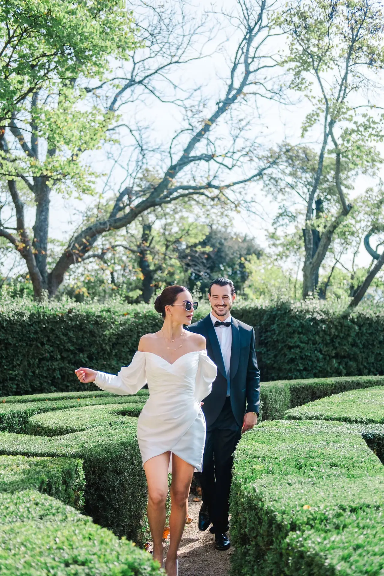 Stylish bride in an off-shoulder satin gown leading her groom through the manicured maze gardens of Château de la Gaude