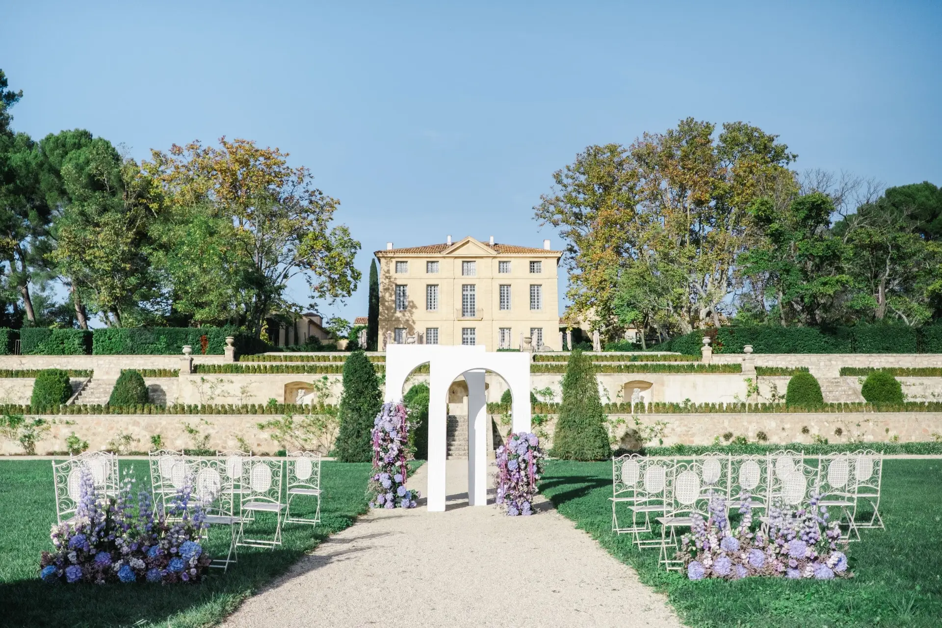 Outdoor wedding ceremony setup with a white arch decorated with purple flowers at Château de la Gaude