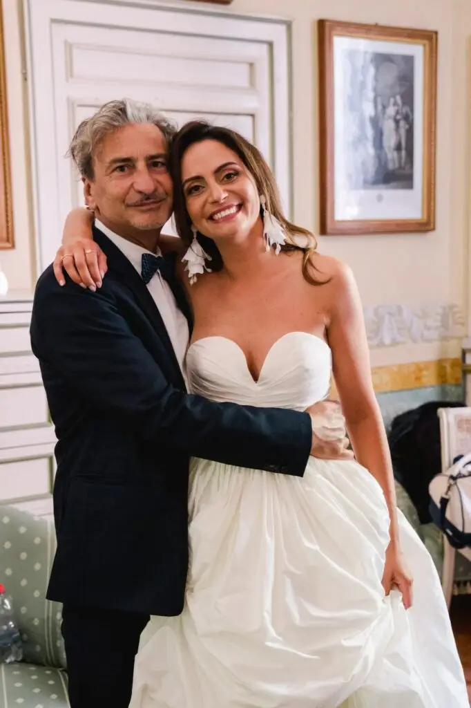 The bride, radiant in white, stands arm-in-arm with her father. Their smiles reflect pride, tenderness, and the anticipation of the ceremony soon to unfold at Villa Pizzo.
