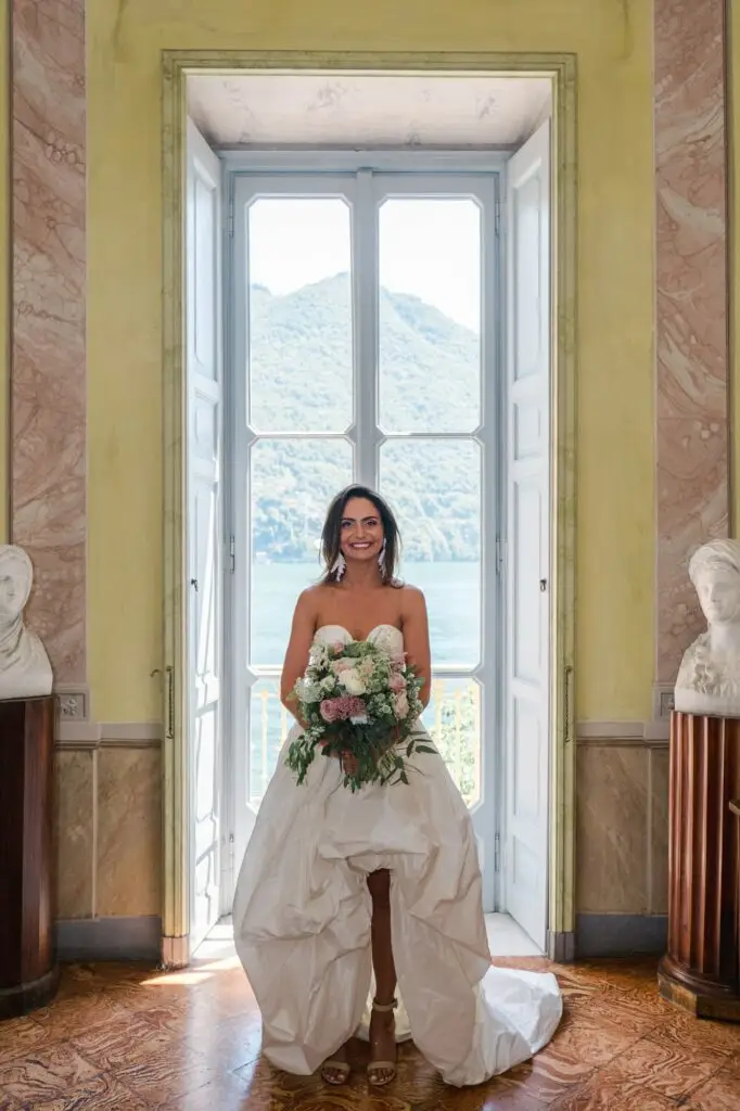 Framed by tall windows overlooking Lake Como, the bride stands poised with her bouquet. The mountains and shimmering waters beyond create a breathtaking backdrop unique to Villa Pizzo.
