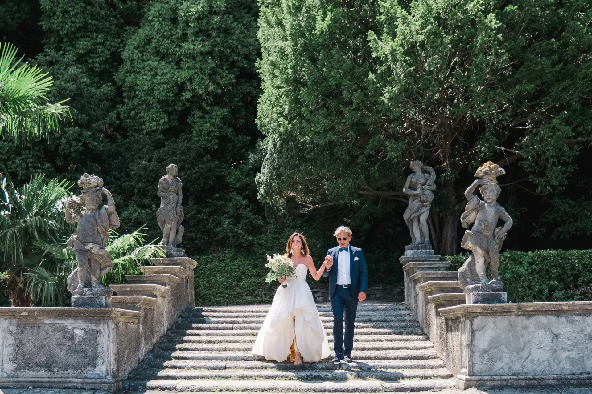 The bride makes her grand entrance, walking hand in hand with her escort down the stone staircase framed by statues.