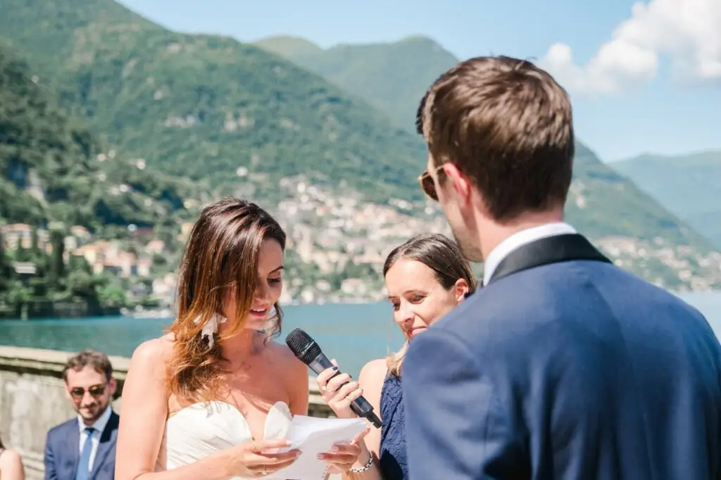 Bride reading her vows with a smile, holding a microphone against the backdrop of Lake Como in Italy.