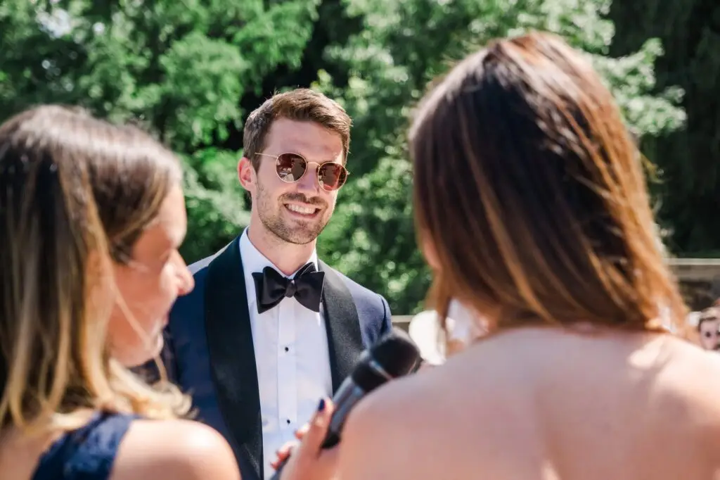 Groom in a navy tuxedo smiling lovingly at his bride while she speaks during the ceremony.
