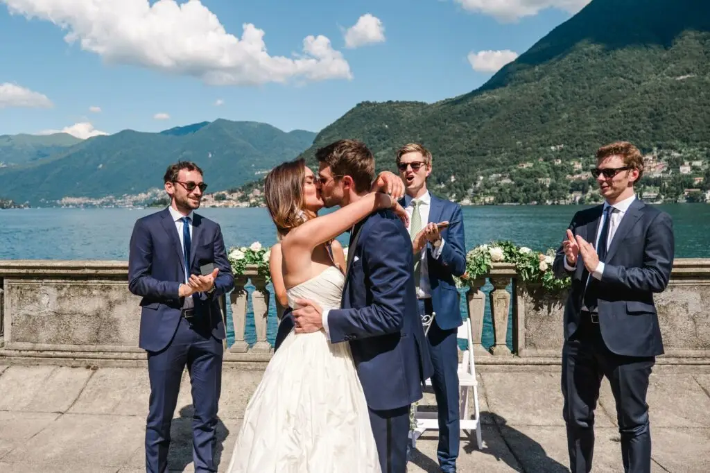 Bride and groom sharing a joyful kiss as guests applaud their union at Villa Pizzo, Italy.