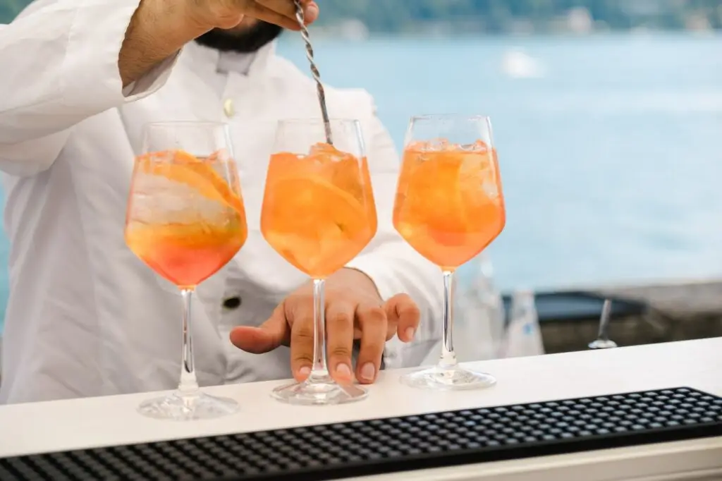 Aperol spritz cocktails being stirred and prepared at a wedding reception bar by the lake.