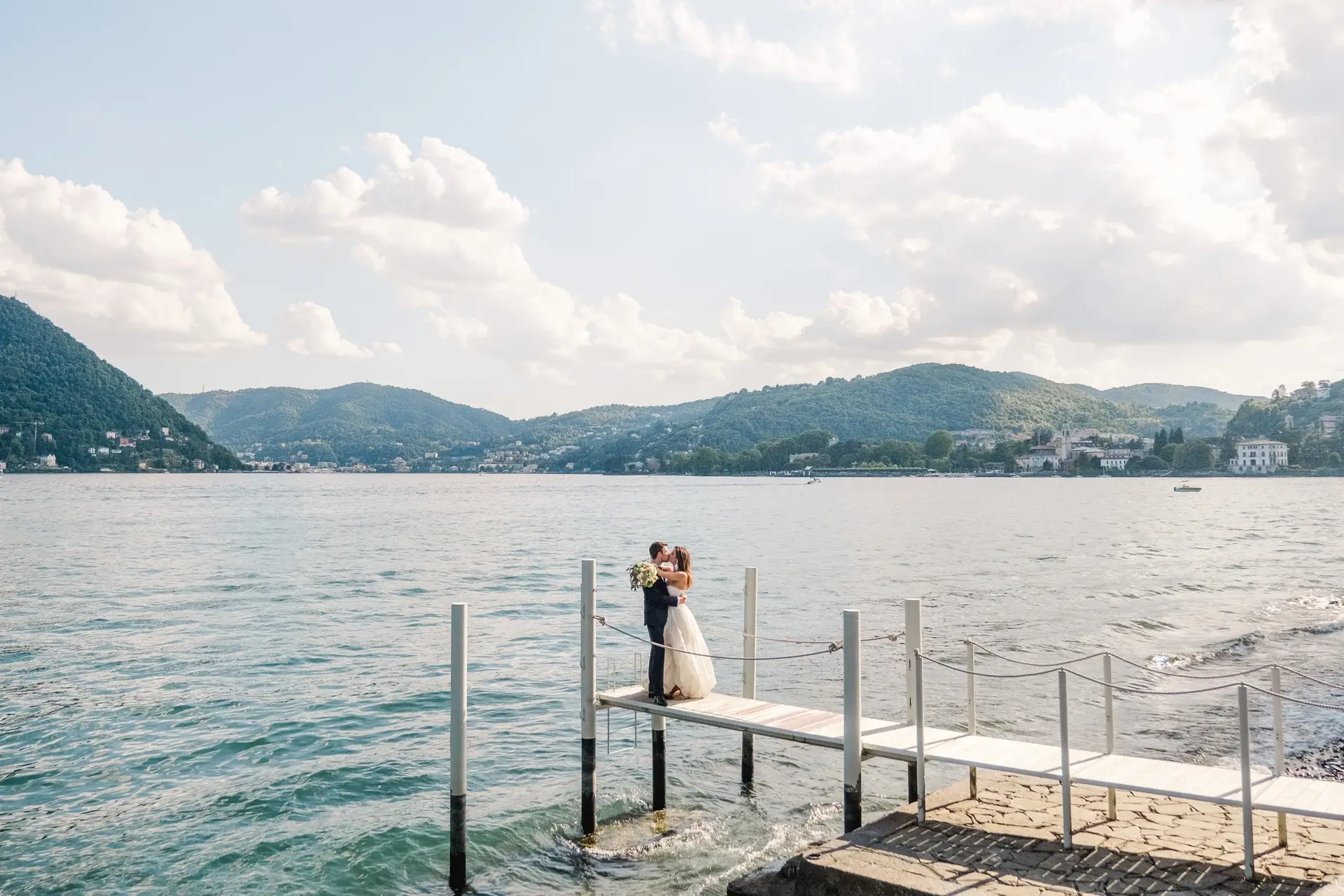 Bride and groom embracing romantically on a wooden pier above Lake Como, surrounded by mountains and blue skies.