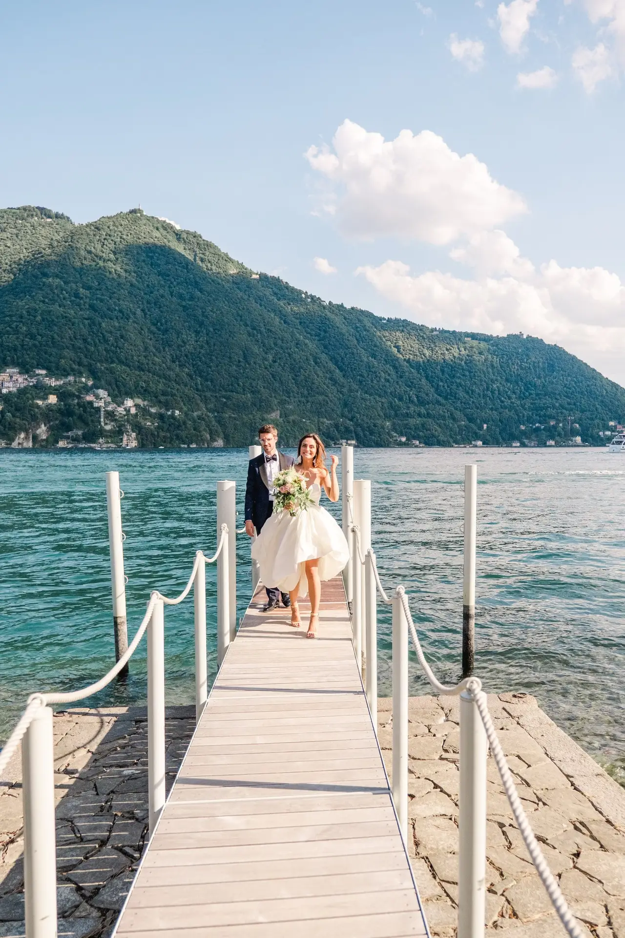 Bride and groom walking barefoot on the dock at Lake Como, with mountains rising in the background.