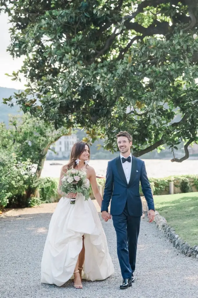 Romantic walk hand in hand through the gardens of Villa Pizzo, the bride holding a bouquet of pastel roses.