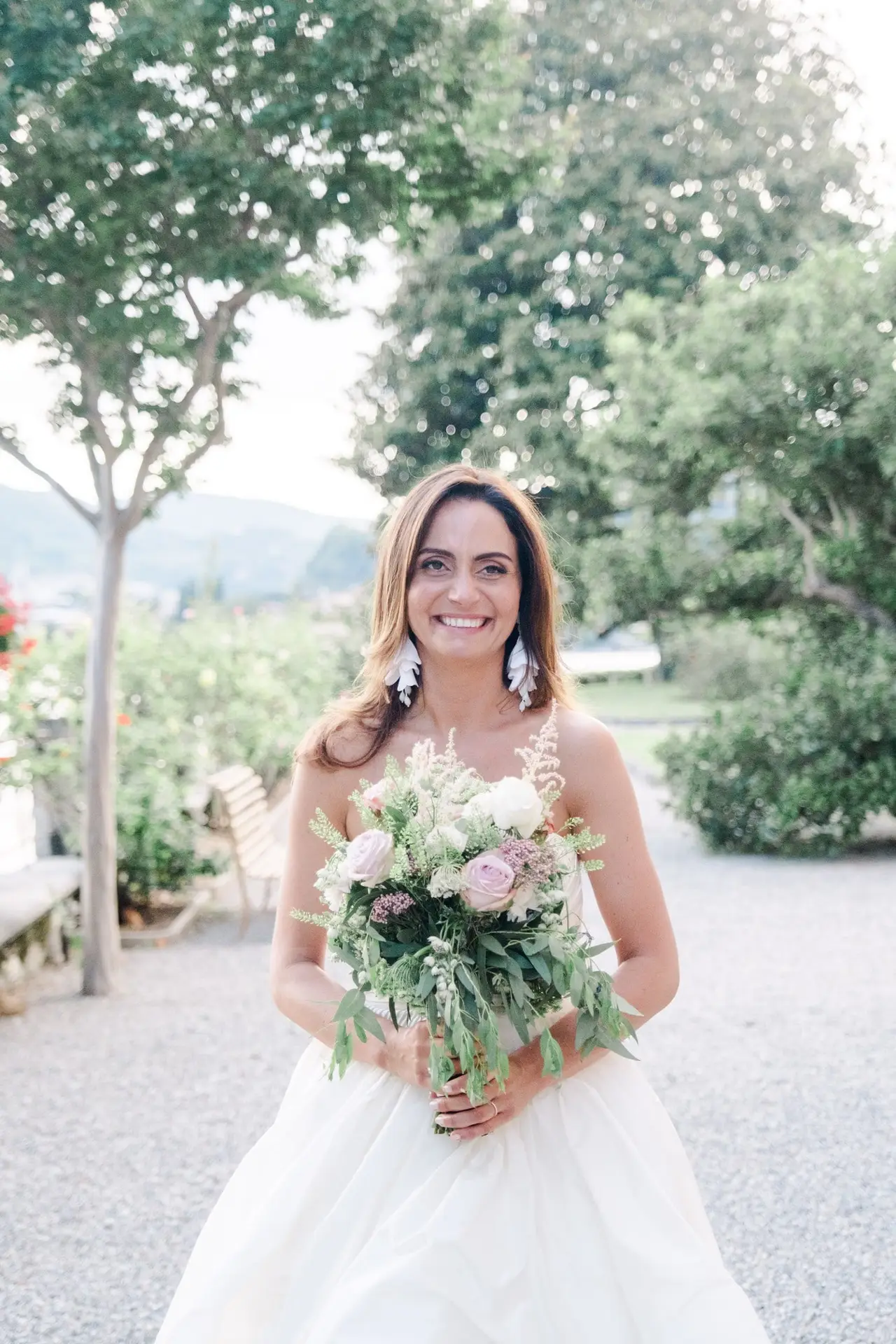 Bride smiling in her white gown, holding a lush bouquet of roses and greenery in the gardens of Villa Pizzo.
