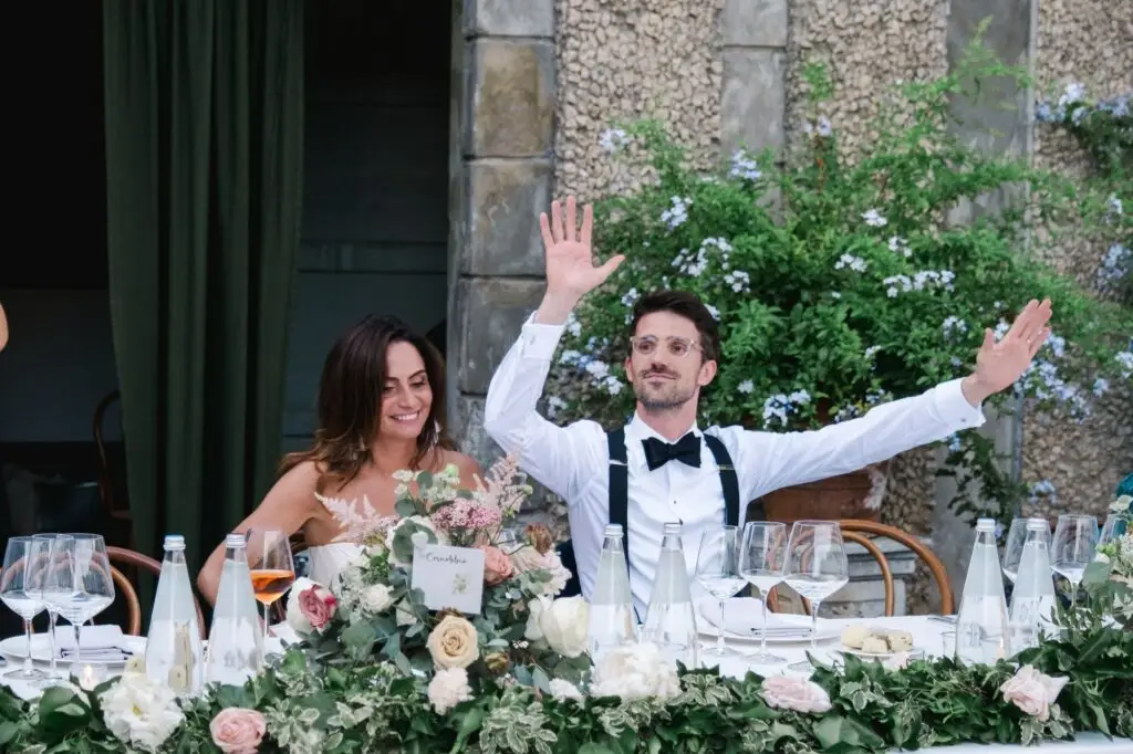 Newlyweds seated at their reception table, smiling and raising their hands in celebration at Lake Como.
