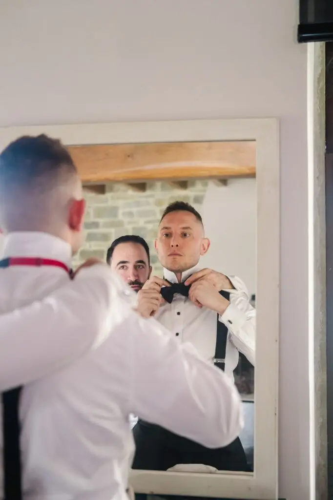 Groom adjusting bow tie in the mirror with help from groomsman during wedding preparations in Tuscany, Italy.