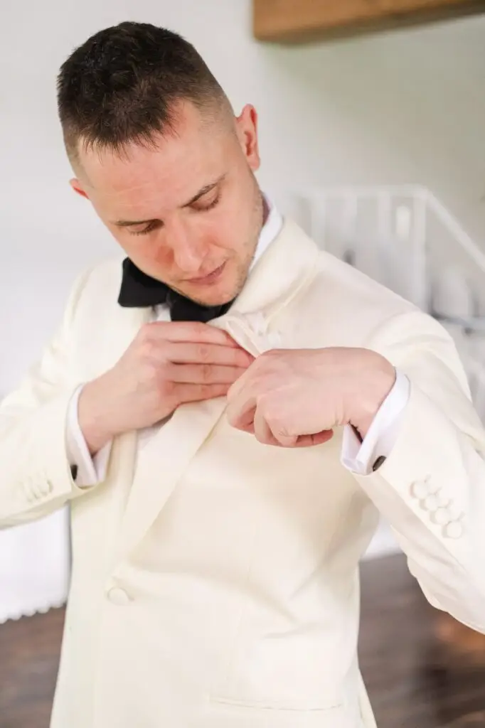 Groom in white tuxedo adjusting boutonniere before wedding ceremony at Borgo Corsignano, Tuscany.