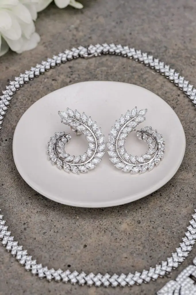 Close-up of sparkling silver earrings with leaf-shaped details, displayed on a small ceramic dish, surrounded by a diamond necklace