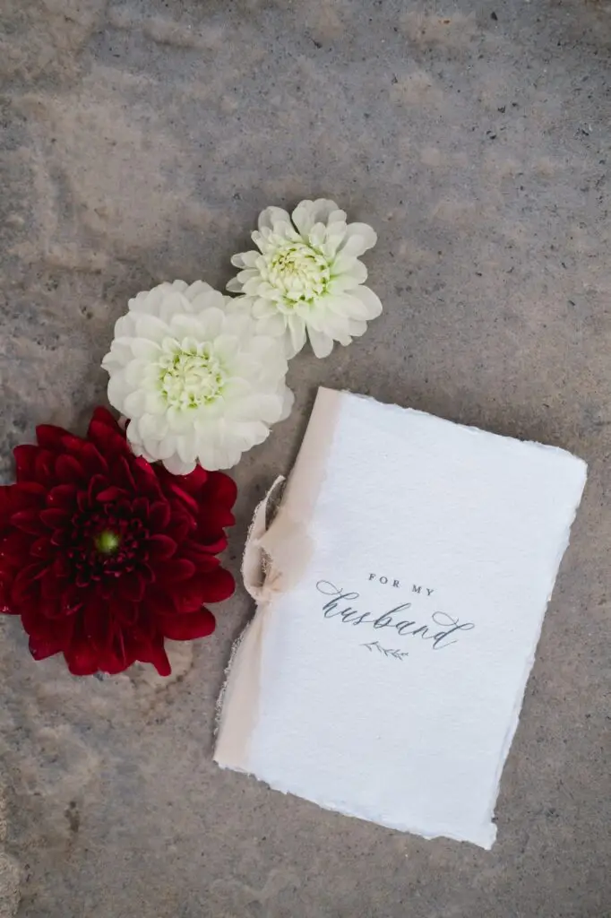 Wedding vow booklet with the words 'For my husband' in elegant script, styled with white and burgundy flowers on stone background