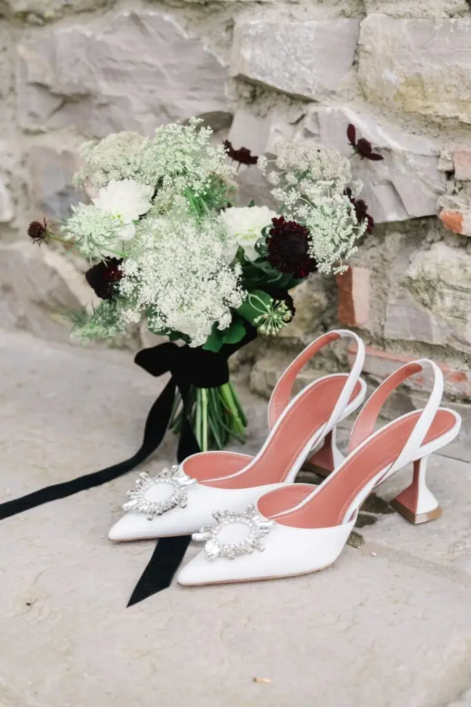 Elegant white bridal heels with jeweled details placed beside a romantic bouquet of white and dark red flowers against a rustic stone wall