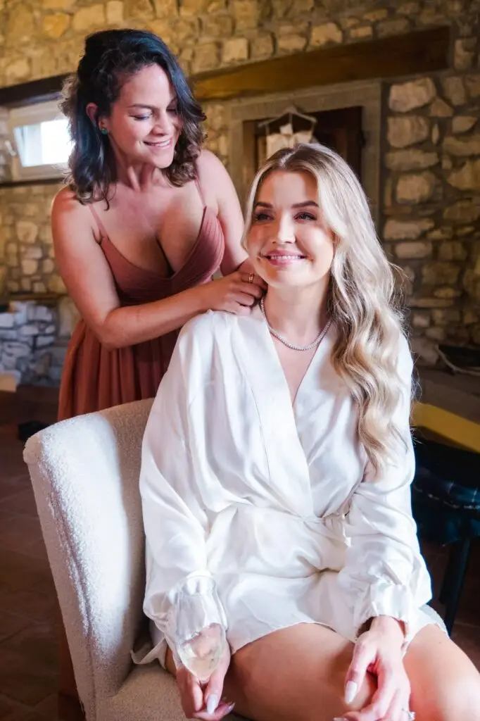 Bridesmaid helping bride put on necklace during wedding preparations in rustic Tuscany venue