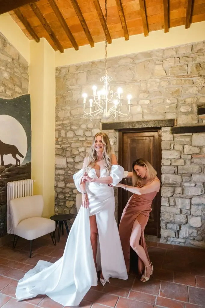 Bride laughing while bridesmaid adjusts her elegant satin wedding gown in a stone-walled Tuscan villa.