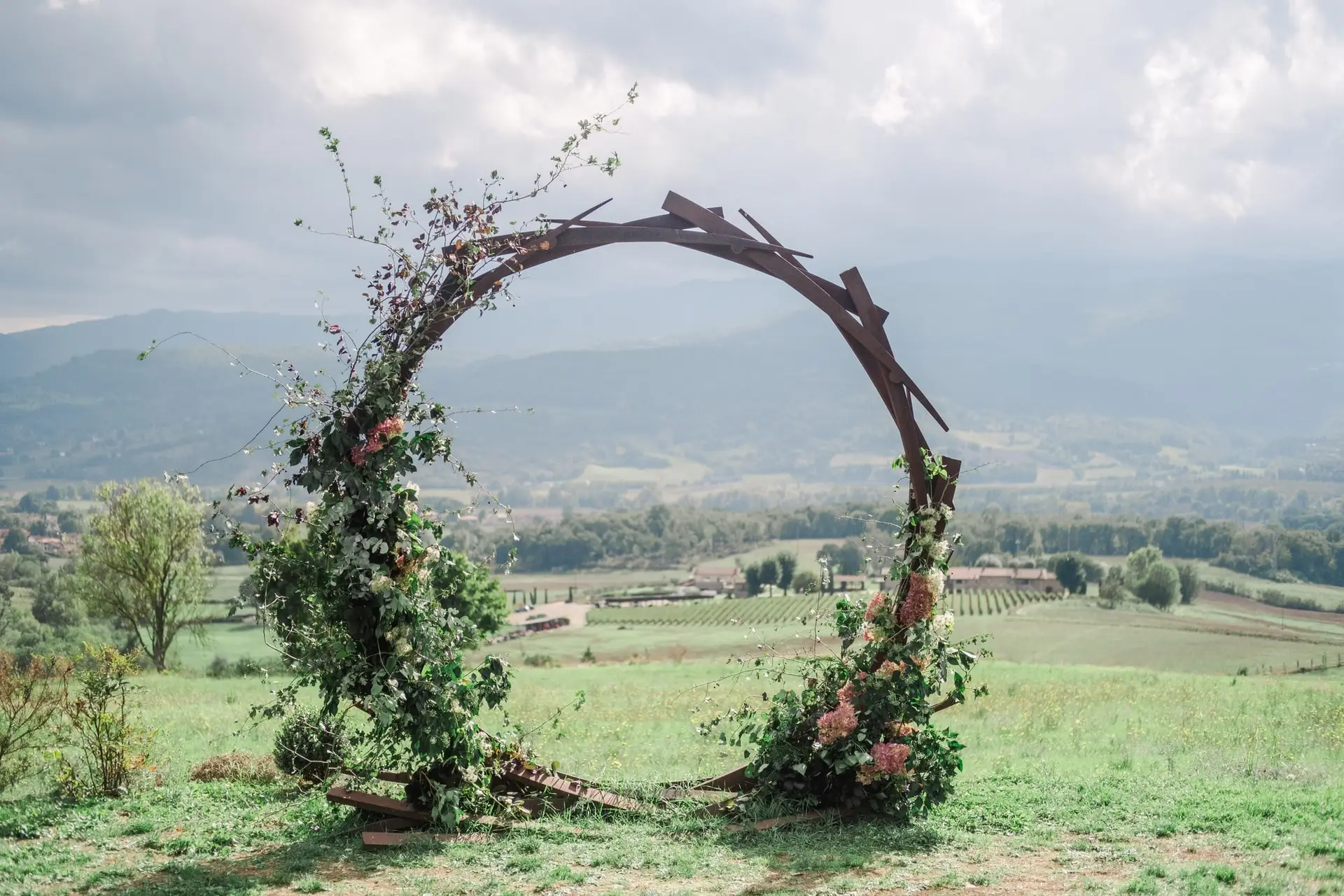 Romantic circular floral arch decorated with vines and blush flowers, overlooking rolling Tuscan hills and vineyards, perfect for a destination wedding ceremony