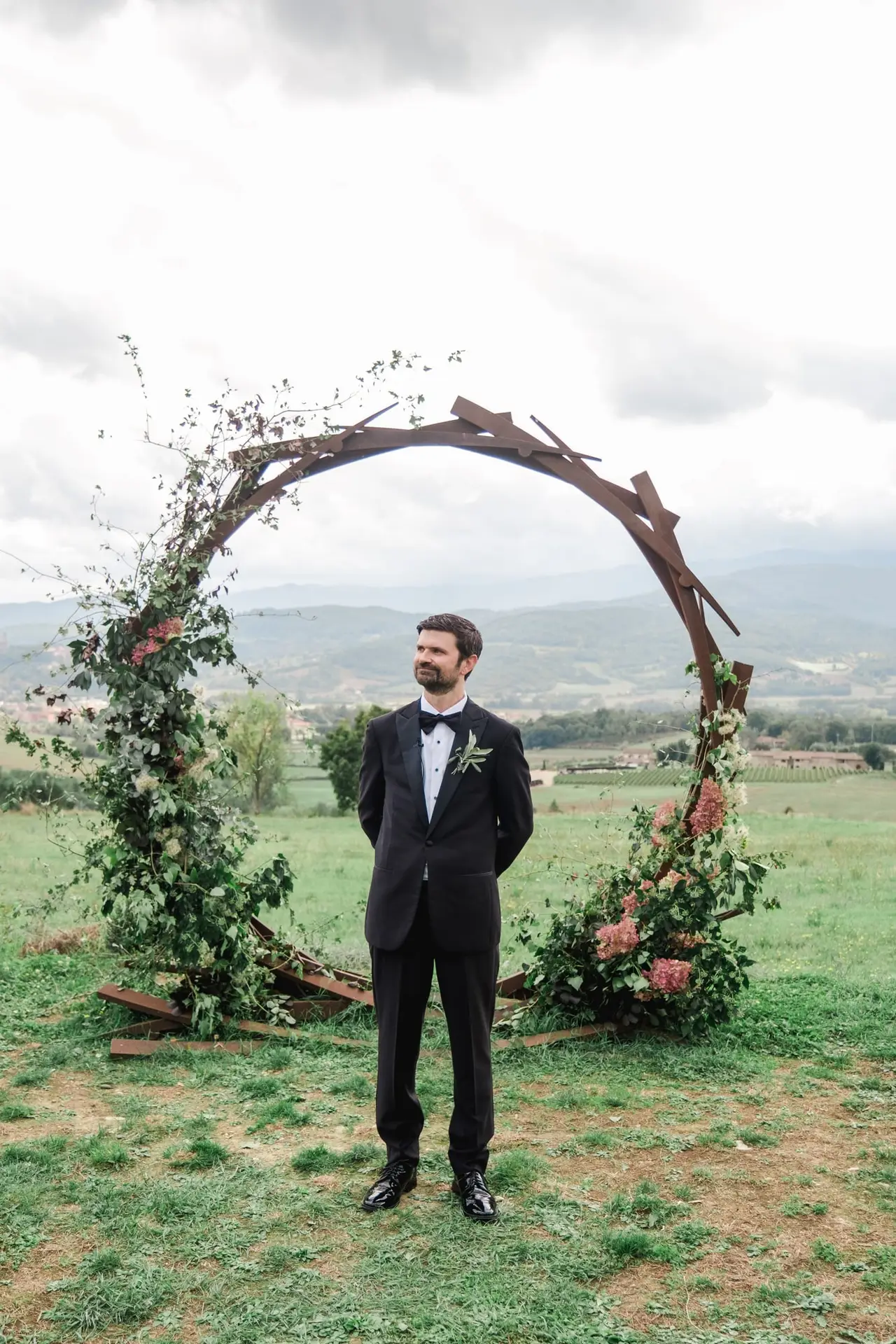 As the Master of Ceremony, I present to you the groom, dressed in a classic black tuxedo, standing beneath a circular floral arch in the heart of the Tuscan countryside, awaiting his bride for the wedding ceremony.