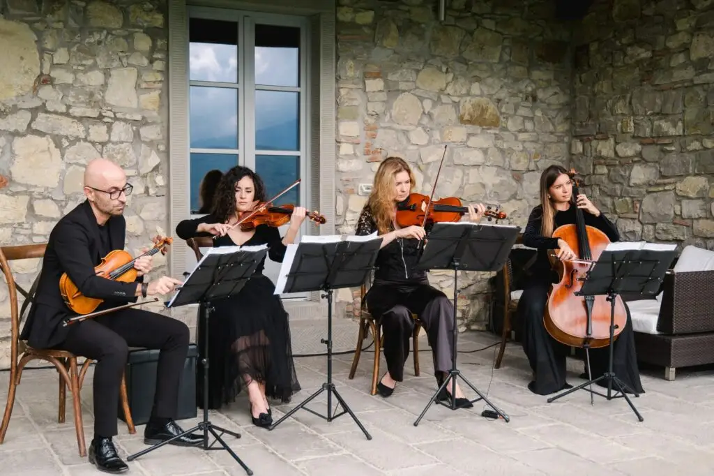 A classical string quartet dressed in black performs outdoors in front of a rustic stone wall, with violins, viola, and cello on display.