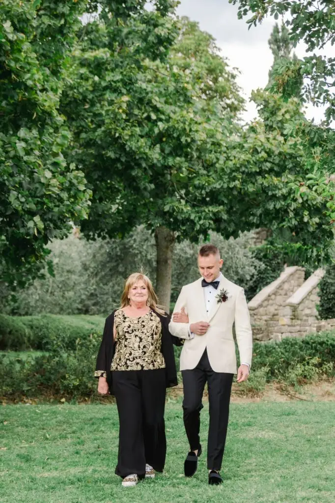 A groom in a white tuxedo jacket walks arm in arm with his mother, who wears a black and gold floral outfit, through a green garden.