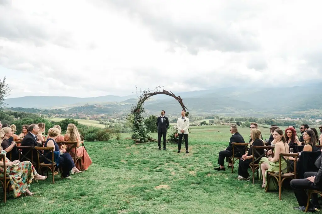Guests are seated on wooden chairs in a lush outdoor ceremony space, with the couple standing beneath a circular floral arch against a Tuscan countryside backdrop.