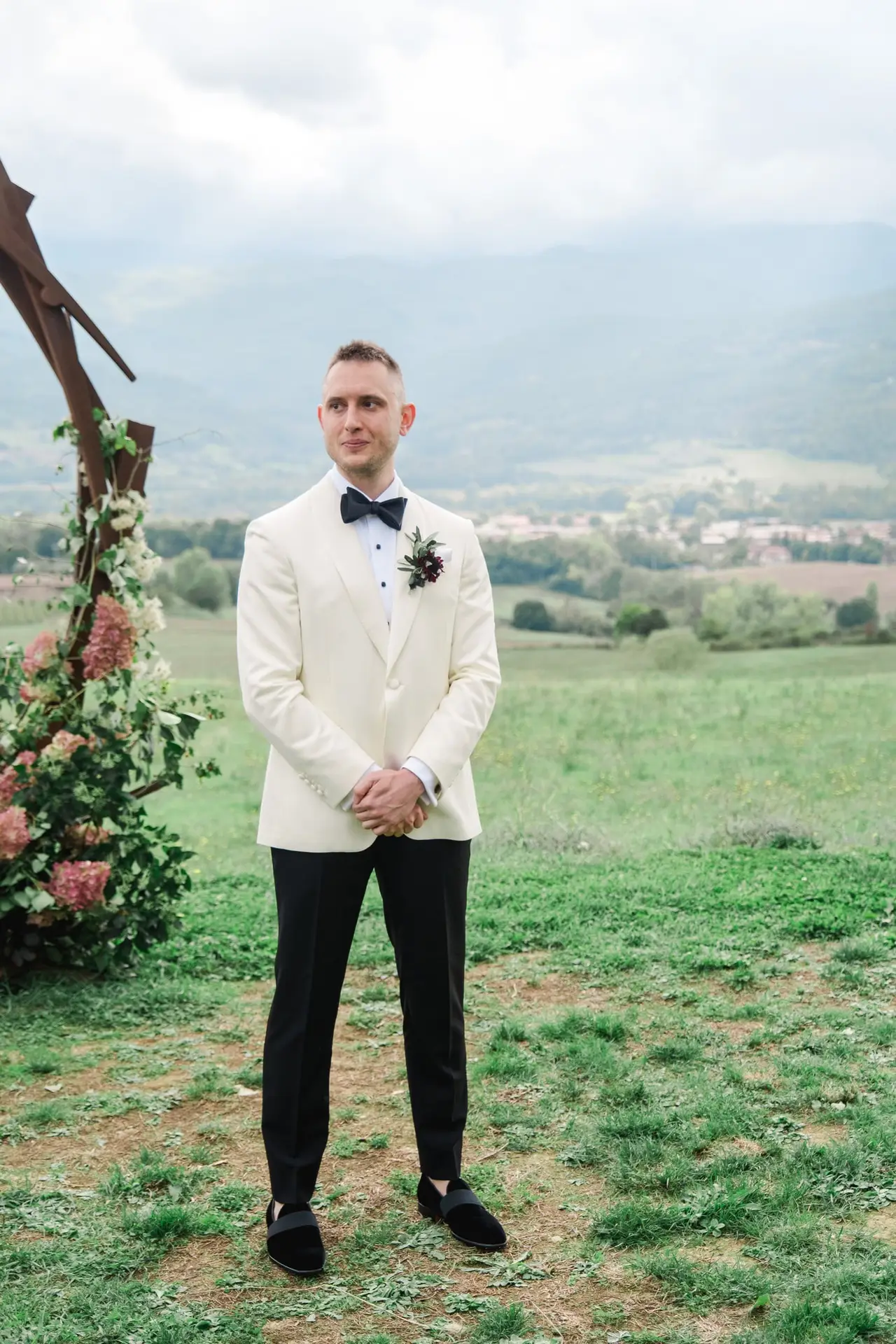 A groom in a white tuxedo jacket and black bow tie stands beneath a floral wedding arch, smiling with rolling hills and vineyards in the background.
