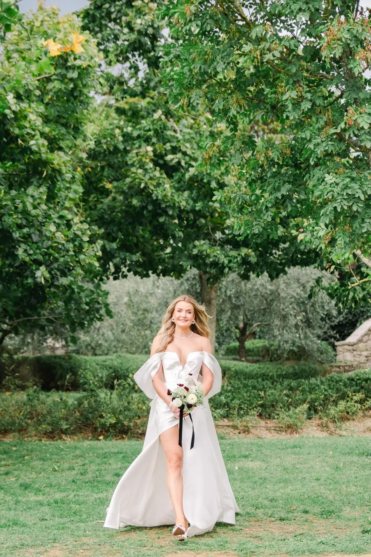 The bride walks gracefully with a bouquet in hand, wearing a luxurious white gown, surrounded by lush greenery in Tuscany.