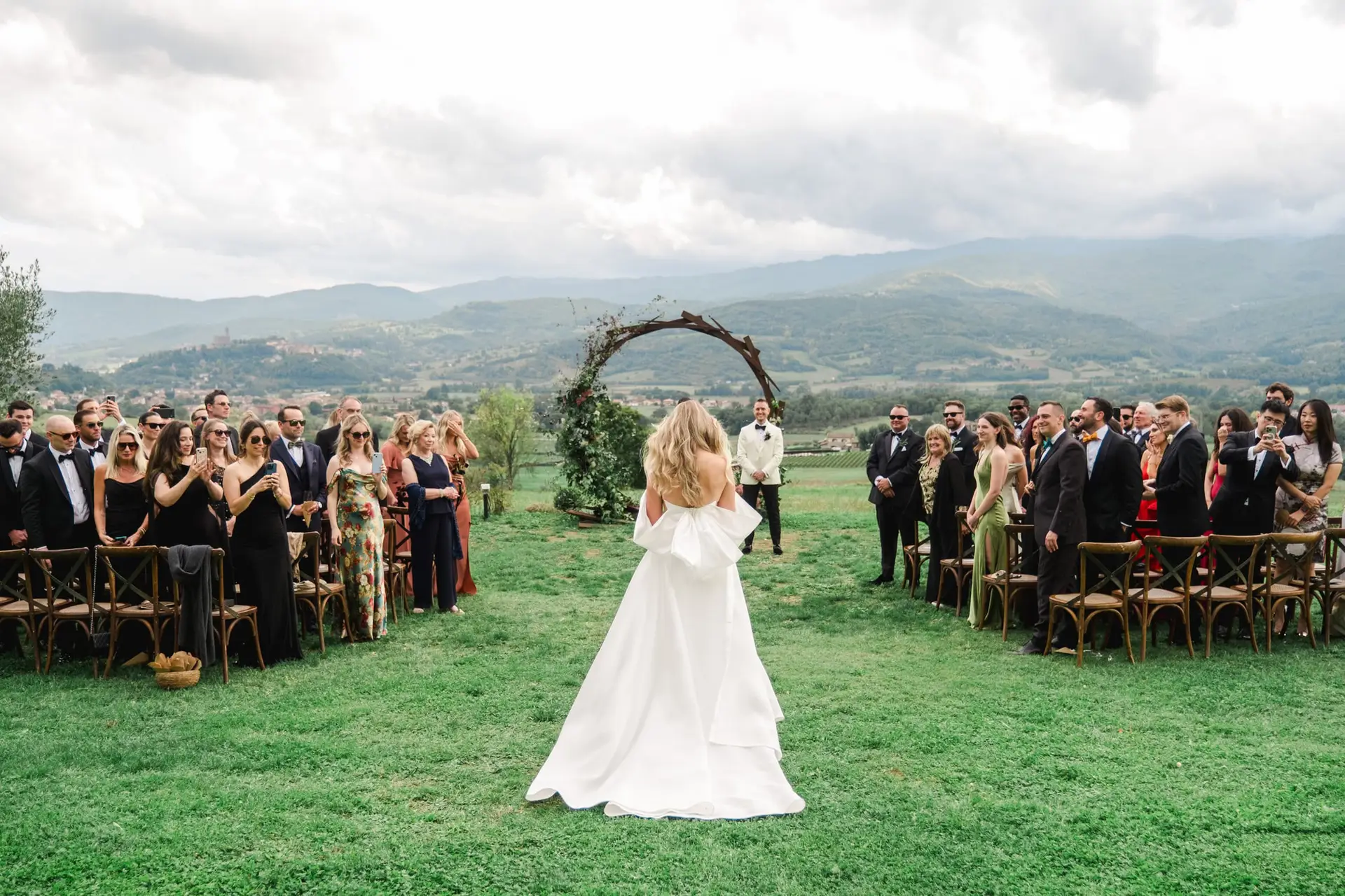 Rear view of the bride walking down the aisle toward the altar, guests standing in admiration, with the Tuscan hills as a backdrop at Borgo di Corsignano.