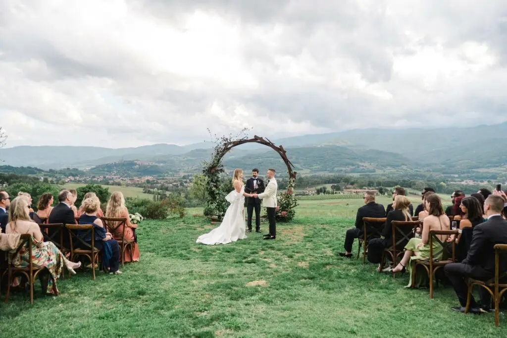 The couple holds hands under a floral arch, exchanging vows in a refined setting overlooking the rolling hills of Tuscany.
