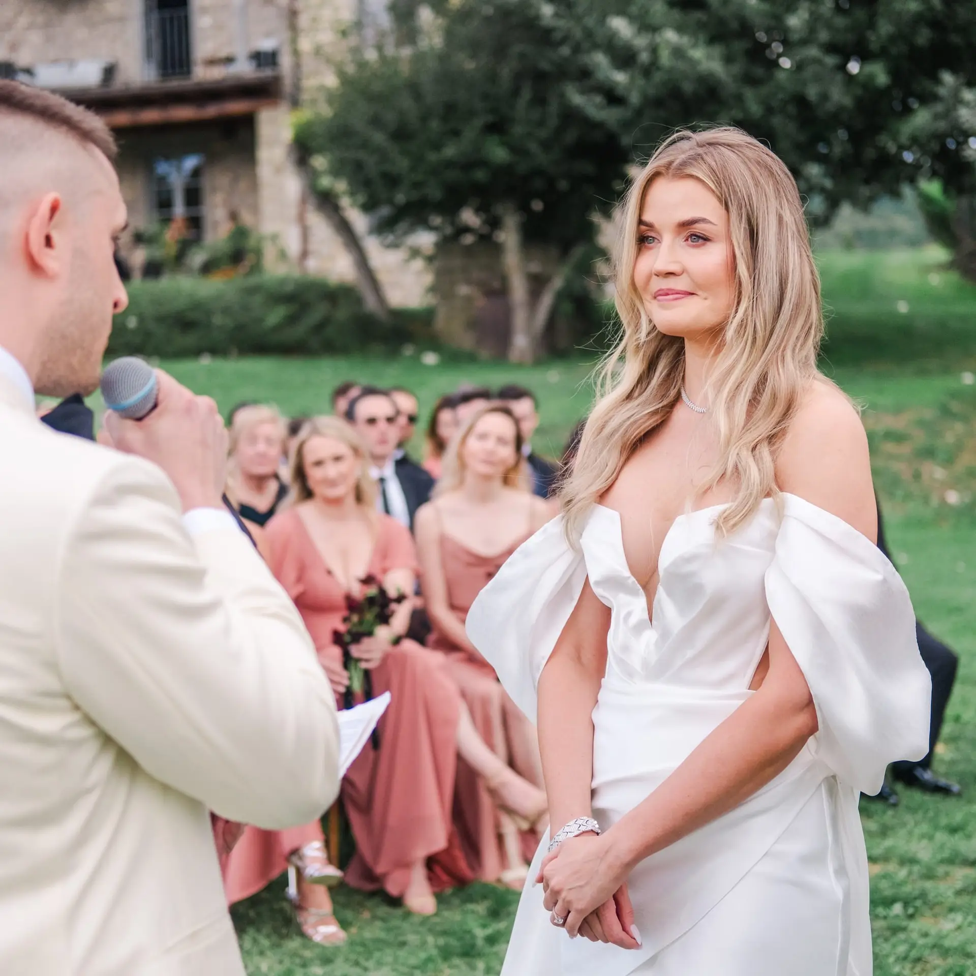 The groom reads heartfelt vows into a microphone, the bride listening attentively, surrounded by their guests at a luxury Tuscan wedding.