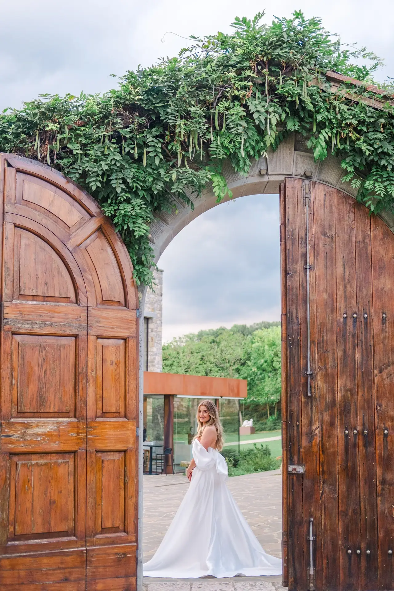 A bride framed by ancient wooden doors and lush greenery at Borgo di Corsignano — elegance and romance captured in one timeless moment, photographed by Julien Boyer.