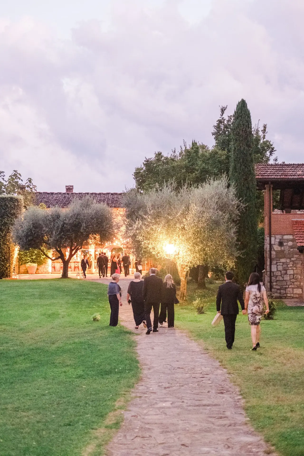 Guests walking along a stone path lined with olive trees towards the illuminated Tuscan villa, creating a refined and welcoming wedding atmosphere, photographed by Julien Boyer.