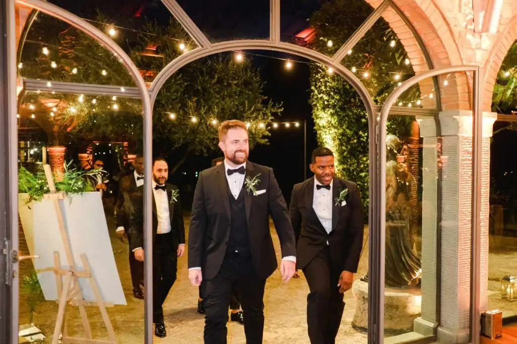 Groomsmen entering the reception hall in black tuxedos, smiling and stylish, under sparkling string lights, photographed by Julien Boyer.