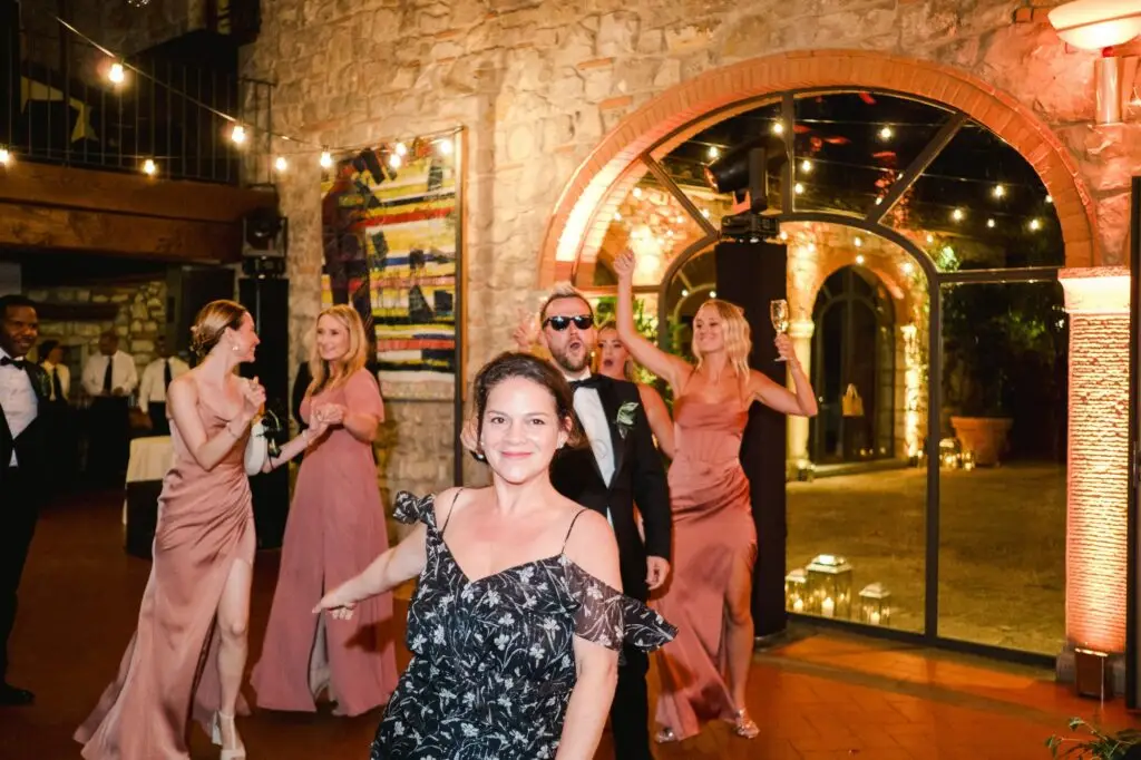 Bridesmaids in satin pink dresses and guests joyfully dancing inside the illuminated stone hall, a festive atmosphere captured by Julien Boyer.