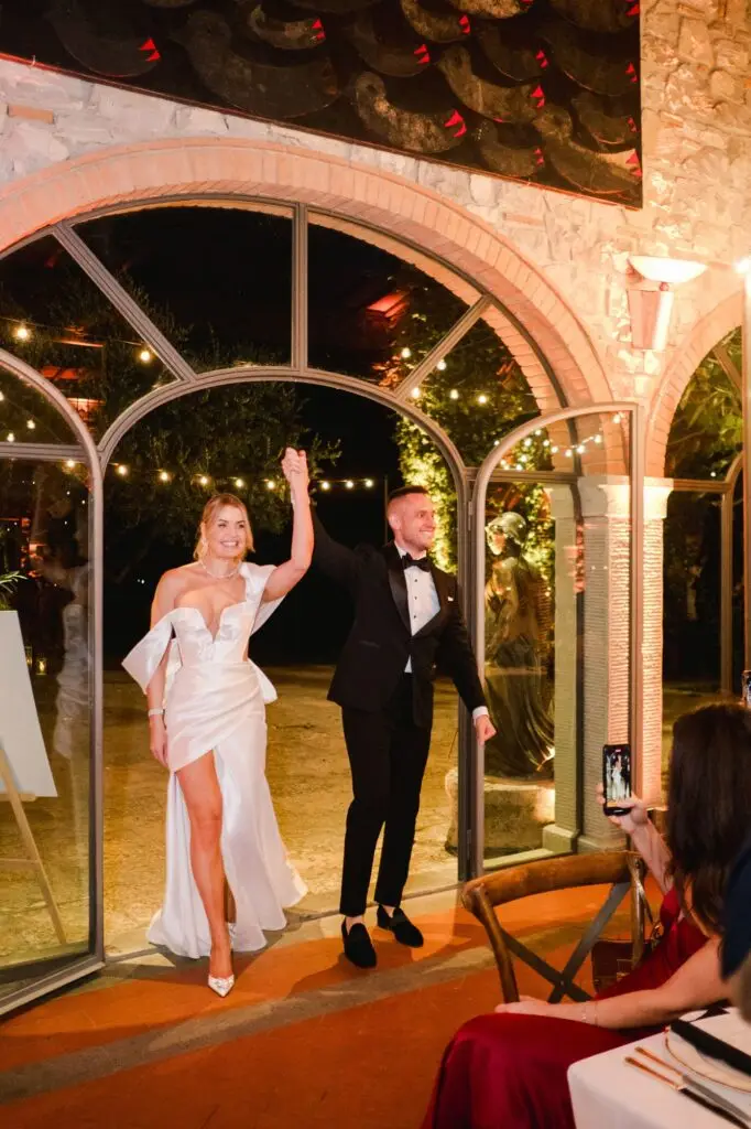 The newlyweds making a grand entrance hand in hand under the glowing arches of Borgo di Corsignano, the bride in a modern and elegant white gown, photographed by Julien Boyer.