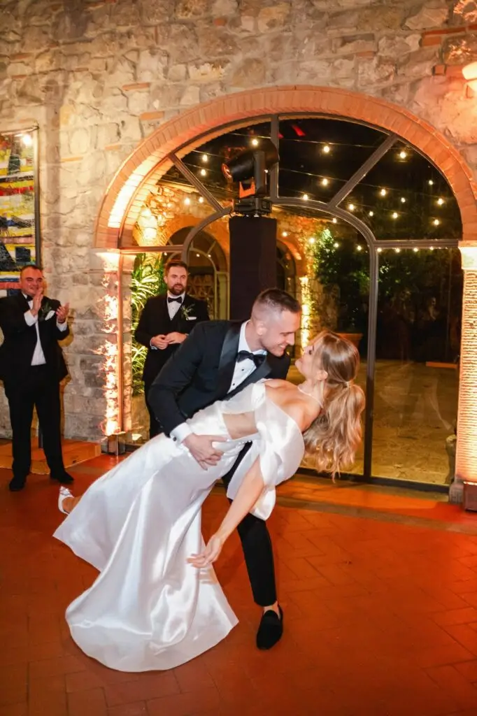 The groom in a black tuxedo dips his bride in her flowing white silk dress for a romantic kiss, as guests applaud around them, captured by Julien Boyer.