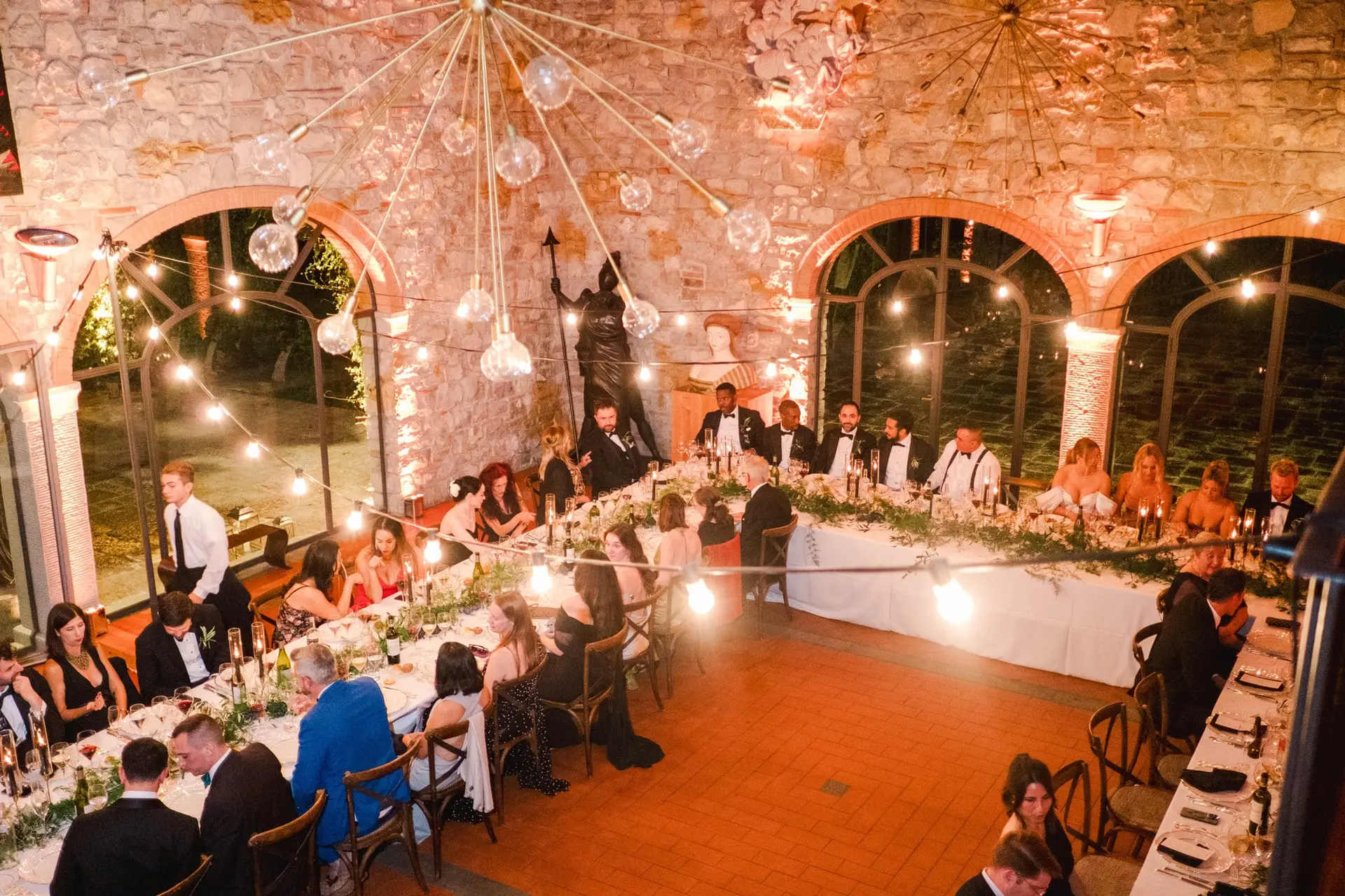 Wide view of the long wedding dinner table inside Borgo di Corsignano, decorated with candles and string lights, surrounded by elegantly dressed guests, photographed by Julien Boyer.
