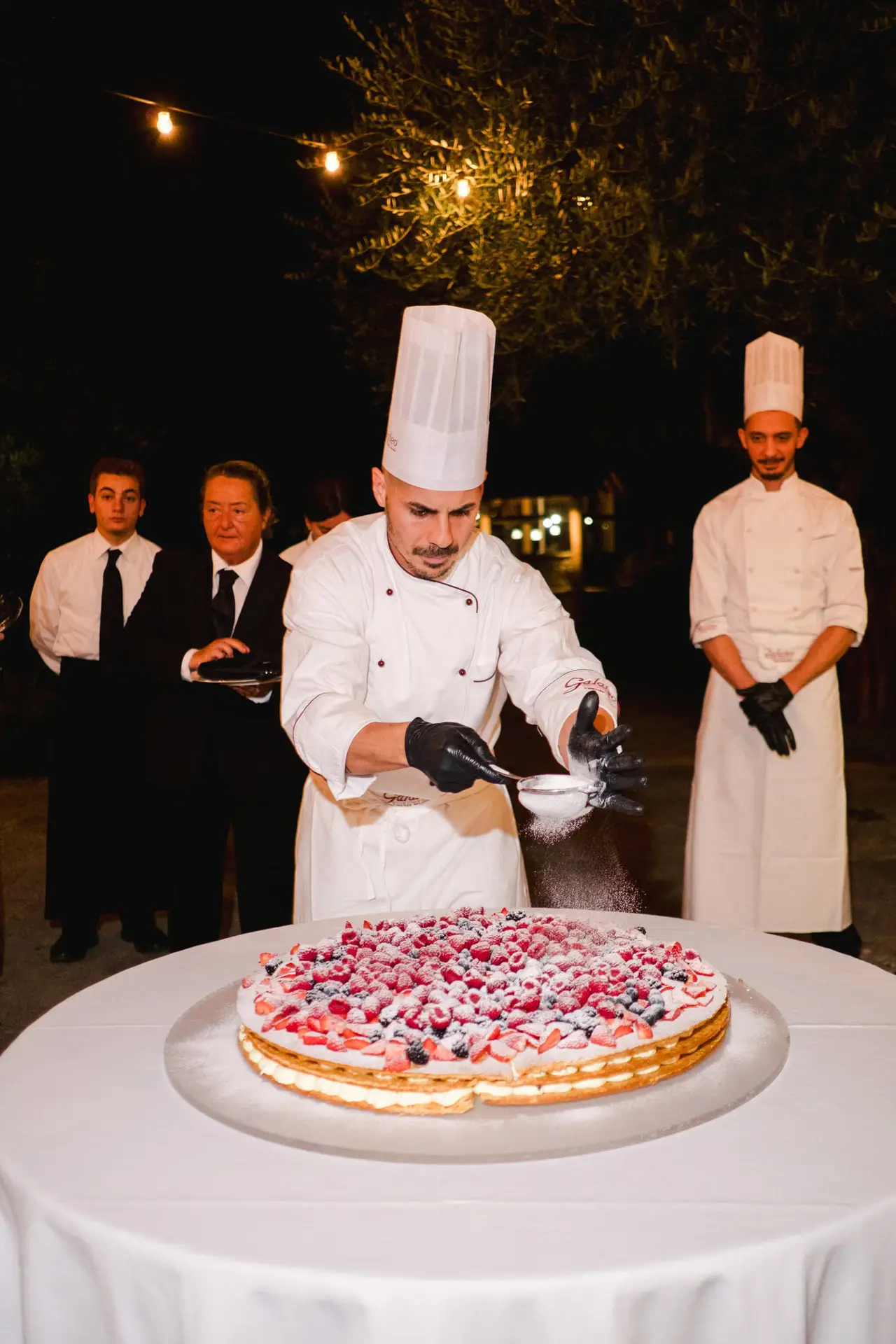 Pastry chef dusting powdered sugar on a large berry wedding cake, surrounded by the catering team during the luxury Tuscan wedding, captured by Julien Boyer.