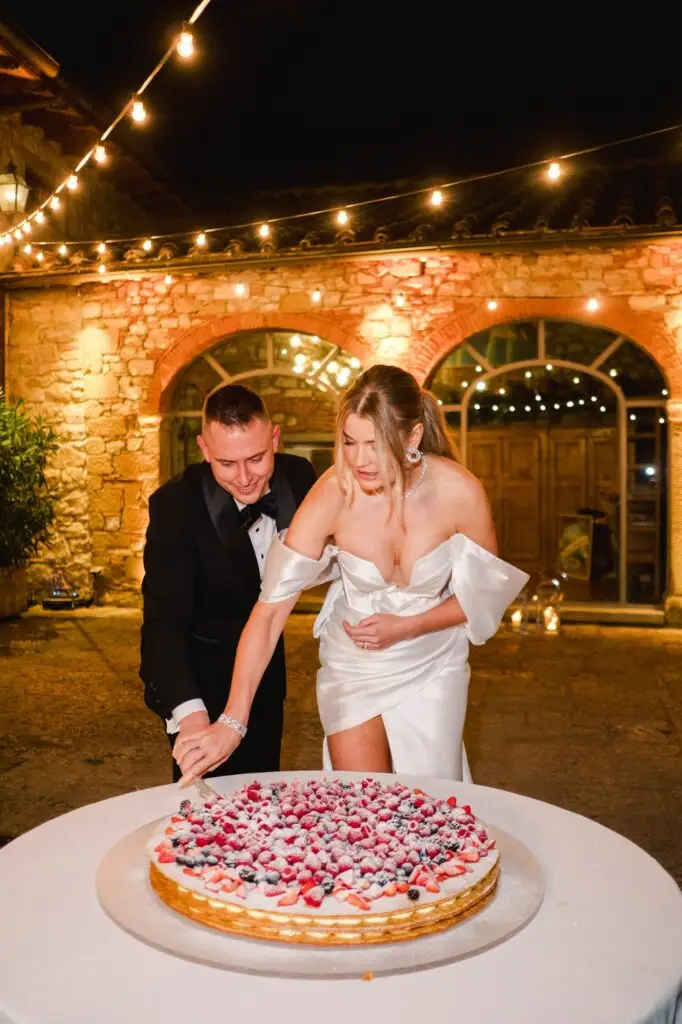 The couple cutting their wedding cake filled with fresh berries under the glowing string lights of Borgo di Corsignano, in a romantic and festive ambiance, photographed by Julien Boyer.