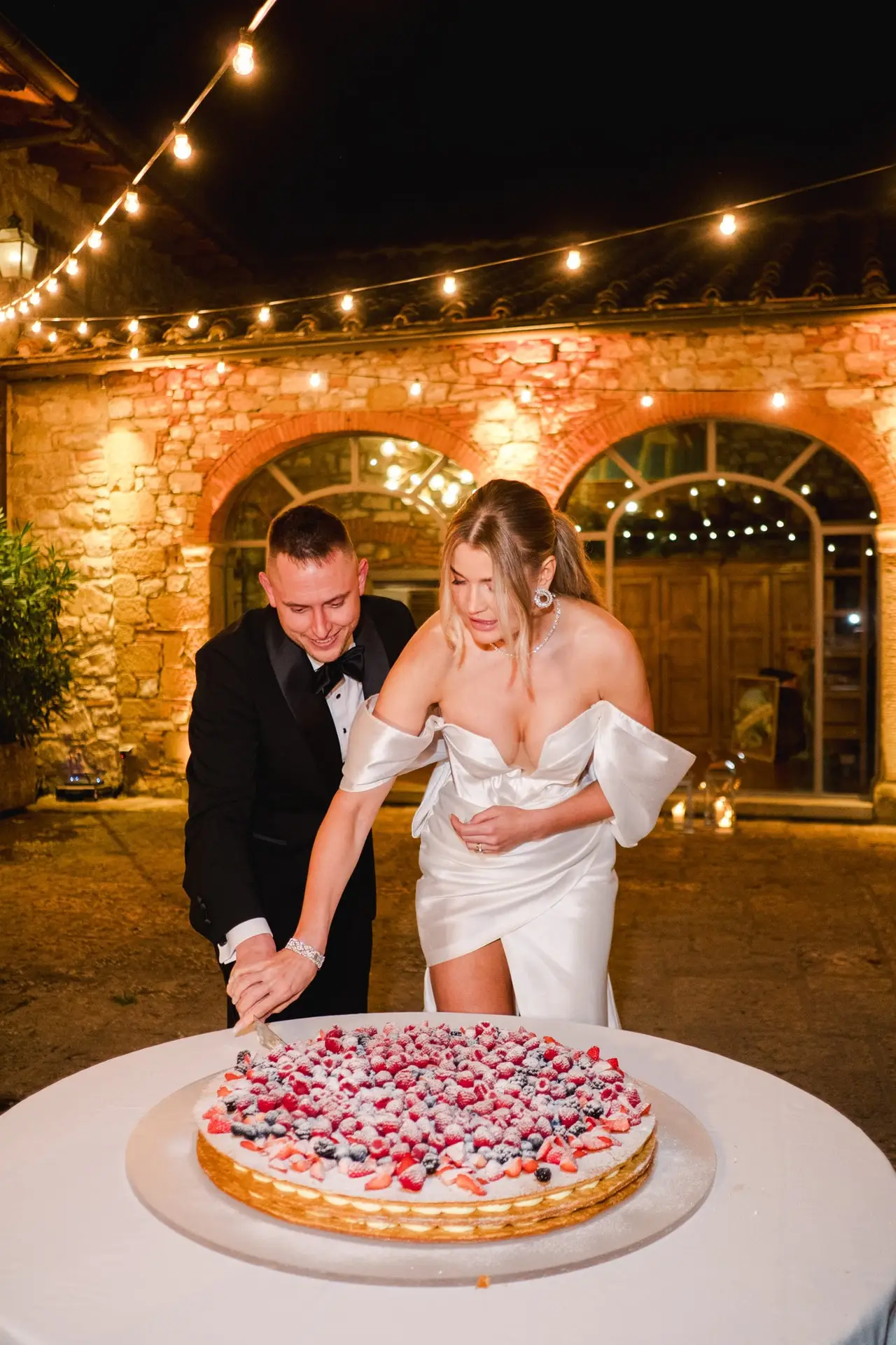 The couple cutting their wedding cake filled with fresh berries under the glowing string lights of Borgo di Corsignano, in a romantic and festive ambiance, photographed by Julien Boyer.